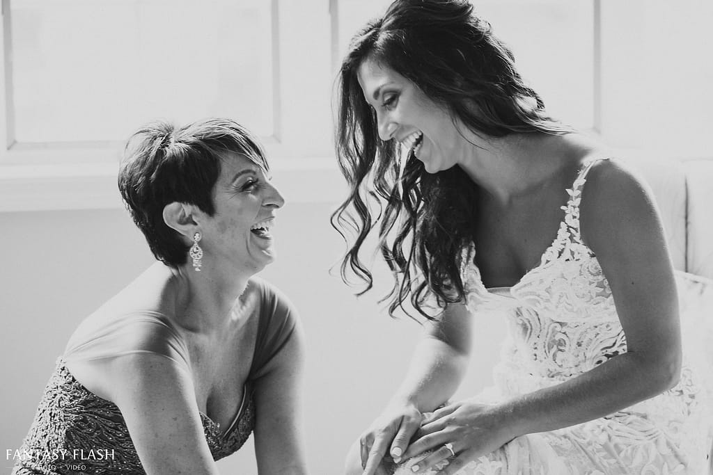This is a black and white photo of a mother and daughter laughing together while getting ready for the daughter's wedding day. The mother is helping the daughter put on her wedding dress.